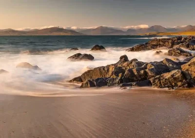 Long-exposure Atlantic waves flowing around coastal rocks with the Harris mountains in the distance.