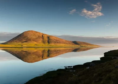 Mountain illuminated by golden evening light and reflected perfectly in calm water in South Harris, Scotland.