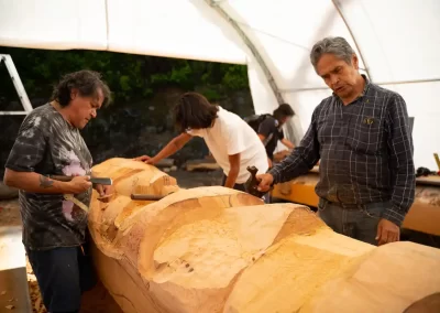 Indigenous woodcarvers shaping a large wooden totem pole beneath a workshop canopy in British Columbia. Photo by Jeremy Williams, courtesy of IISAAK OLAM Foundation.