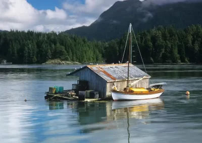 Small sailing boat moored beside a floating wooden building beneath forested mountains near Tofino, photographed by Garth Lenz.