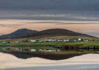 White croft houses and rounded hills reflected in calm coastal water in South Harris, Scotland.