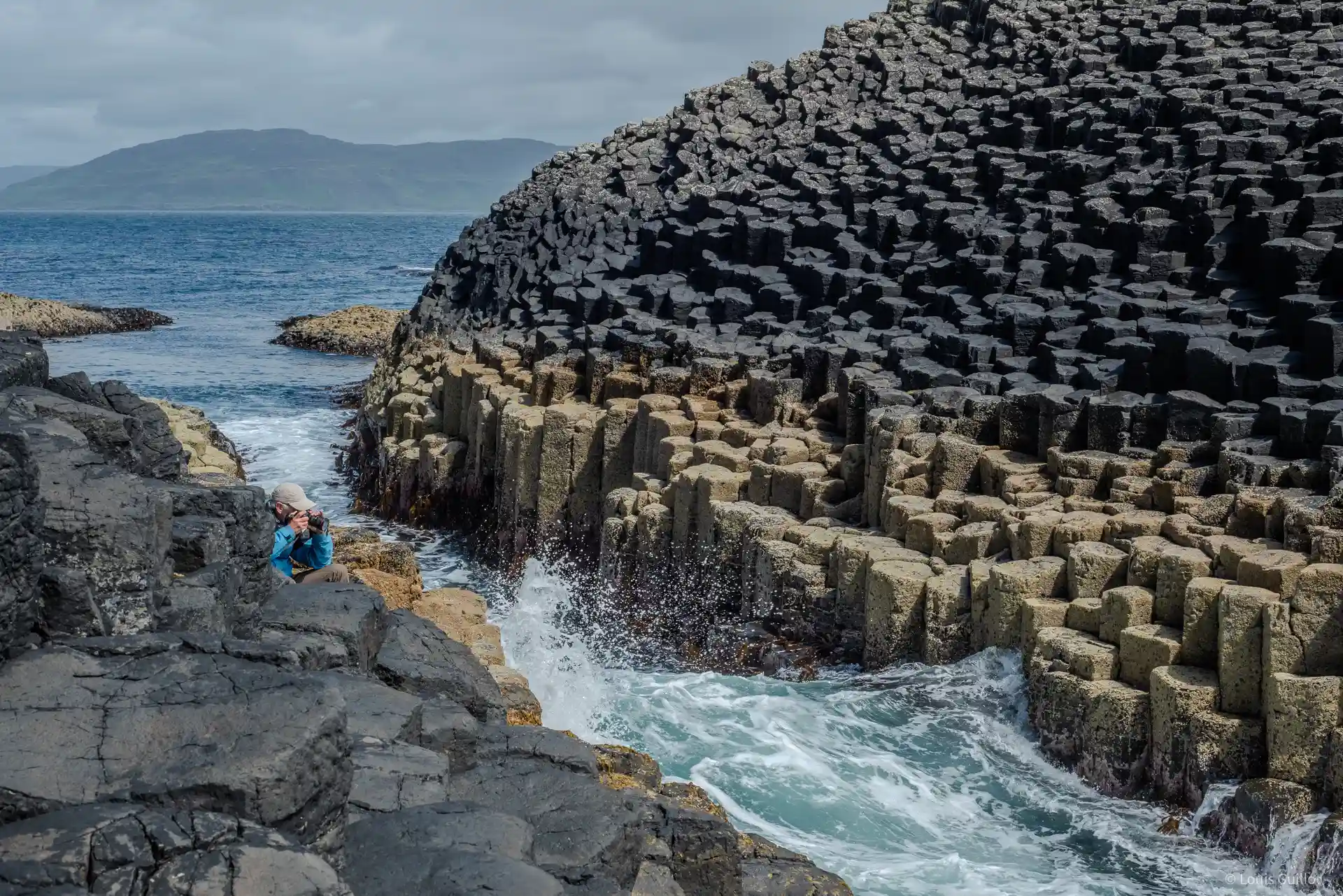 The incredible basalt columns found on Staffa on the west coast of scotland near the isle of mull can be seen here as waves go through a corridor between the basalt columns. A photographer takes photos as the waves crash on these columns.