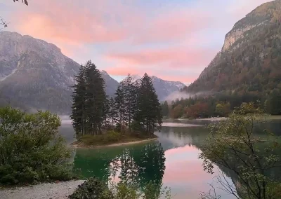 Pink sunrise reflected in a calm alpine lake surrounded by forested mountains in Slovenia.