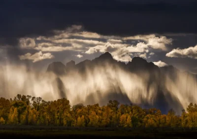 Sunlit curtains of rain sweep across jagged mountains above a line of golden autumn trees.